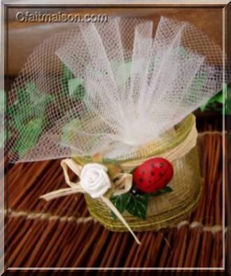 Présentation de dragées dans un pot de laitage en verre bas et droit recouvert d'un ruban rustique, finition avec noeud en raphia naturel, lierre, coccinelle et fleurs.
