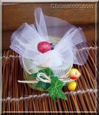 Présentation de dragées dans un pot de laitage en verre bas et droit recouvert d'un ruban rustique, finition avec noeud en raphia naturel, lierre et petites pommes décoratives.
